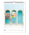 Die Pickmotion Zauberhafte Welt - Kalender (30x40) zeigt einen Elefanten, der mit seinem Rüssel bunte Luftballons hält und zwischen weißen Bögen vor einer blauen Wand steht.