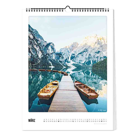 Das Bild "Der Berg ruft - Kalender" (30x40) von Pickmotion zeigt hölzerne Anlegestellen mit Ruderbooten auf einem ruhigen Bergsee, umringt von schneebedeckten Gipfeln im Vorfrühling.