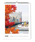 Eine Uferszene mit herbstlichen Bäumen, einem angedockten Boot und modernen Gebäuden - abgebildet im Hamburg - Kalender (Format 30x40) von Pickmotion, ideal für jeden Fotokalender mit nordischem Design.