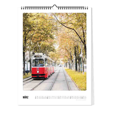 Auf der Seite Pickmotion Wien - Kalender (Format 30x40), März, fährt eine rote Straßenbahn an herbstlichen Bäumen vorbei und fängt die prächtige Architektur und den lebendigen Charme Wiens in atemberaubenden großformatigen Bildern ein.